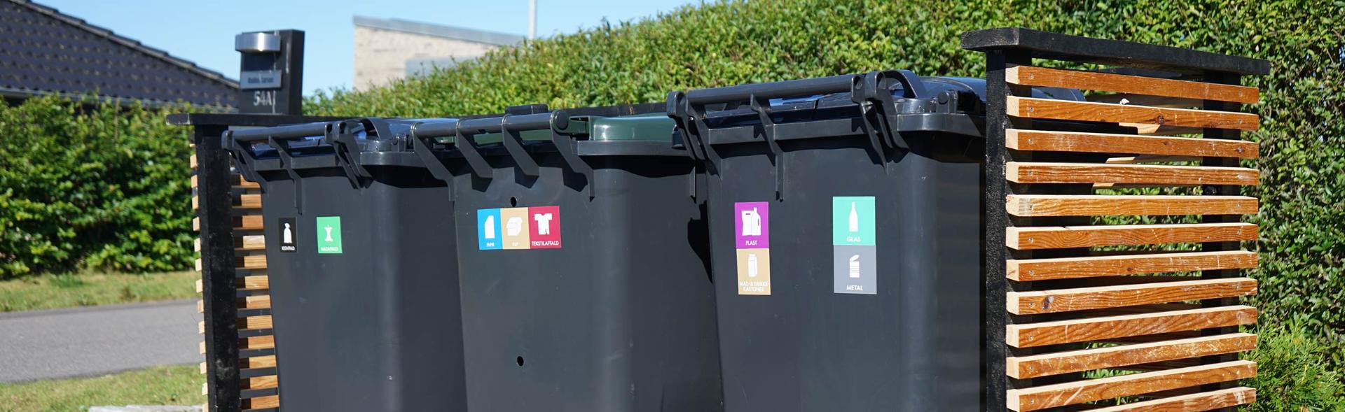 Tre affaldsbeholdere står på række i skjul langs hæk